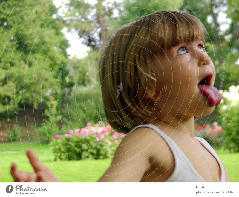 Funny, funny, cheerful, happy, funny blonde girl outside in the garden, looks up and sticks out her red tongue. Little joker is fooling around, fun with twisted eyes in nature, in the park with flowers and trees.