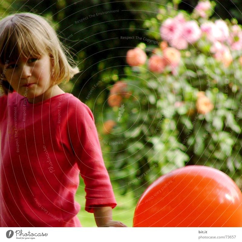 Blonde, sweet girl is standing with a red balloon in her hand in the garden in front of pink flowers in full bloom. Birthday child outside in the nature is happy about the balloon for her party.